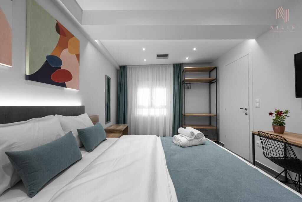 A modern hotel room with a neatly made bed, a desk with a chair, a wardrobe, and a large abstract painting on the wall.