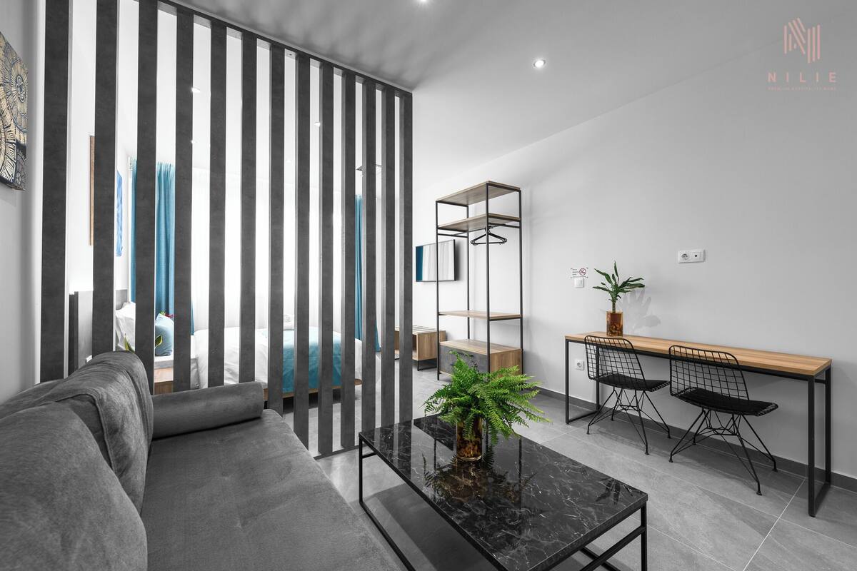 Un salon moderne comprenant un canapé, une table basse avec une plante, un bureau avec deux chaises et une porte coulissante avec des stores verticaux.