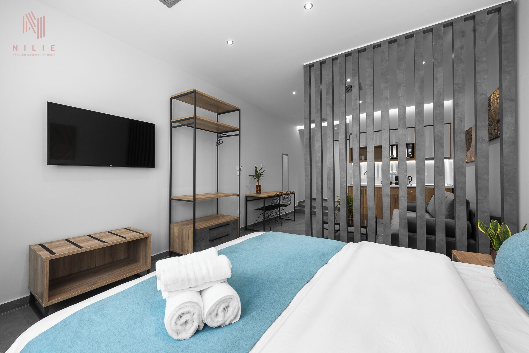 A modern hotel room with a bed, a TV, a wooden shelf, a desk, and a grey partition.