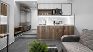 A modern kitchen with wooden cabinets, a built-in microwave, and a coffee maker. There is a grey sofa and a plant in the living area. - Factory Inn Nilie Hospitality MGMT (Thessaloniki)