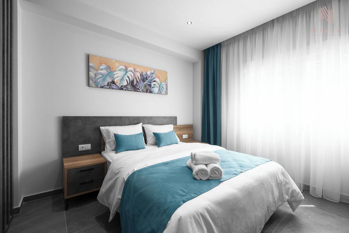 Une chambre d’hôtel moderne avec un lit bien fait, une table de chevet, un tableau au mur et une grande fenêtre avec des rideaux.