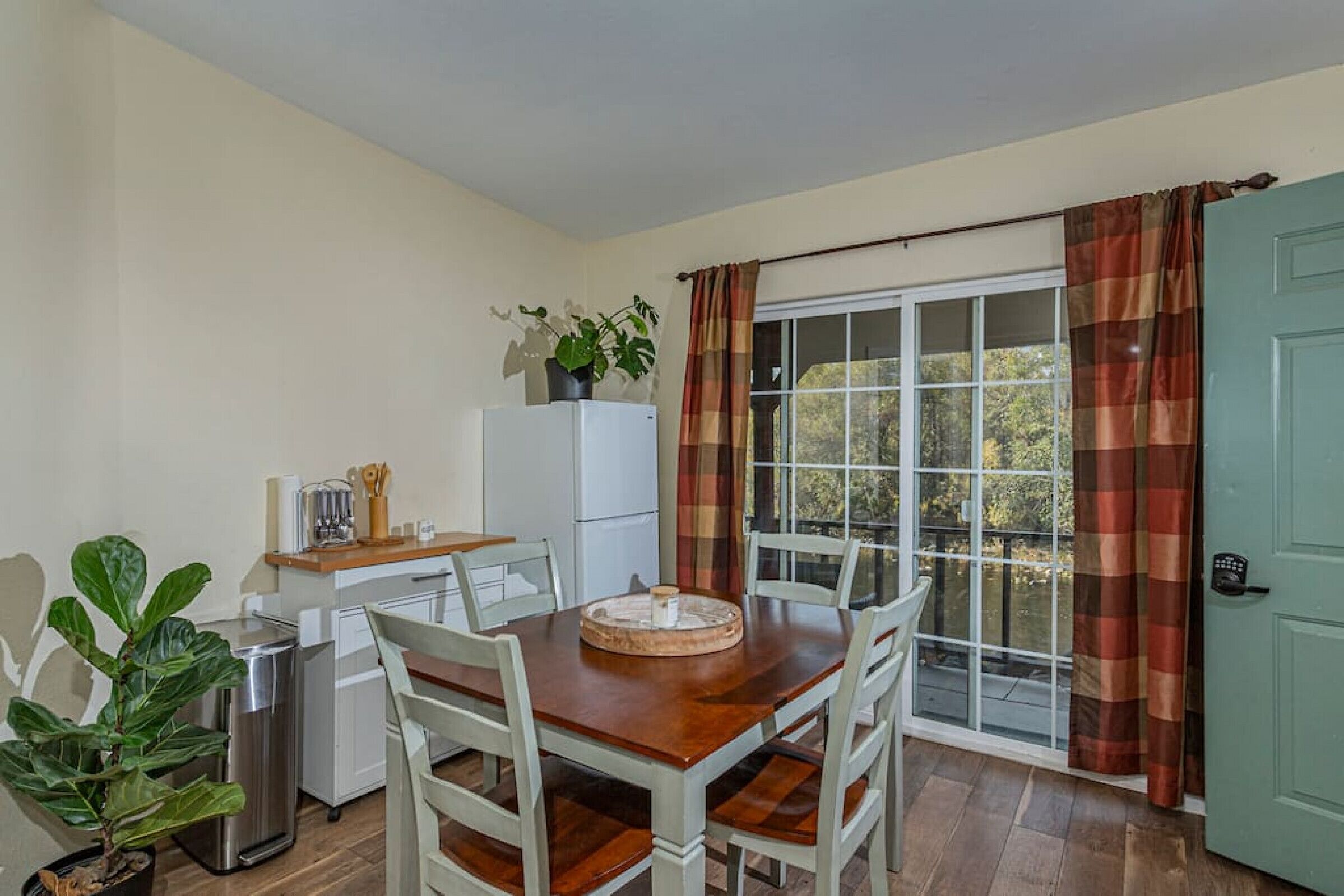 A dining area with a wooden table and white chairs, a refrigerator, a potted plant, and a window with curtains.