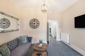Ein Wohnzimmer mit einer Couch, zwei Uhren, einer Pflanze auf einem Couchtisch, einem Fernseher an der Wand und einer Tür zu einem anderen Raum.