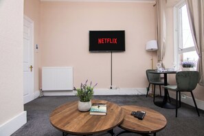 Ein Zimmer mit einem Fernseher, der 'NETFLIX' zeigt, einem runden Holztisch mit einem Buch und einer Fernbedienung, einer Topfpflanze und einem Fenster mit Vorhängen.