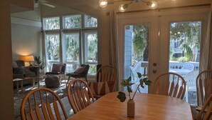 Ein Essbereich mit Holztisch, Stühlen und einer Vase mit Pflanze. Große Fenster mit Vorhängen, ein Deckenventilator und ein Blick ins Freie.