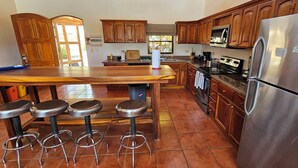 A kitchen with wooden cabinets, a stainless steel refrigerator, a microwave, and a wooden bar stool area. - Great House (Guanacaste Province)