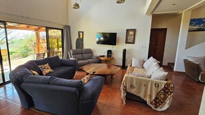 A spacious living area with a sofa, armchair, and a coffee table. A flat-screen TV is mounted on the wall, and there is a sliding glass door leading to an outdoor area. - Great House (Guanacaste Province)