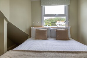 A bedroom with a bed, two bedside lamps, a window with a view of houses and greenery, and a staircase to the left. - Shippen (Kingsbridge)