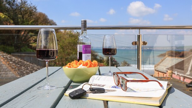 Una mesa con dos copas de vino, una botella, un bol de aperitivos, un par de gafas, un libro y llaves en un balcón con vistas panorámicas.