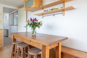 A wooden dining table with three stools, a vase with flowers, and wooden shelves above holding glasses and mugs. - Shippen (Kingsbridge)