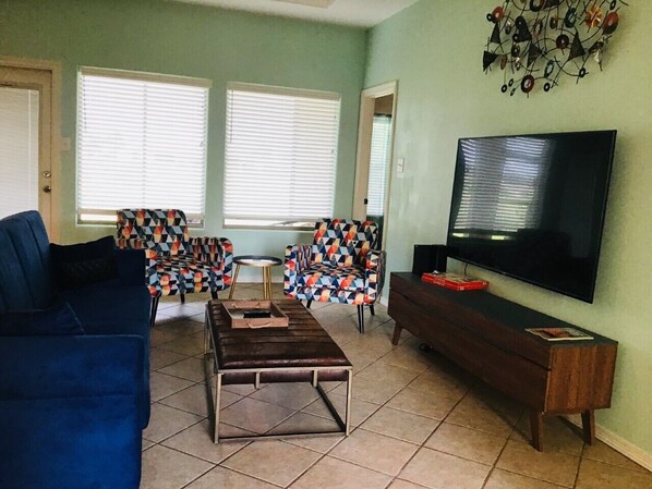 Uma sala de estar com um sofá azul, cadeiras estampadas, uma mesa de centro, uma TV e um armário de madeira.