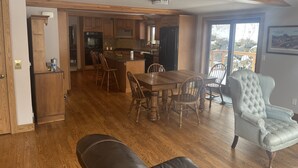 A spacious living area with a wooden floor, a dining table with chairs, a leather sofa, and a large window offering a view of the outdoors.