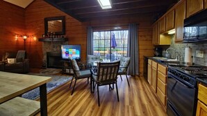 A cozy cabin interior with a wooden interior, a fireplace, a dining area with chairs, a kitchen with a stove and cabinets, and a TV displaying a streaming service interface. - Cozy Cabin~Sleeps 6~Great Location~Dog-Friendly (Branson)