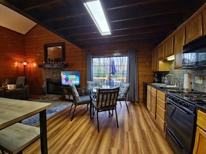 A cozy cabin interior with a wooden interior, a fireplace, a dining area with chairs, a kitchen with a stove and cabinets, and a TV displaying a streaming service interface.