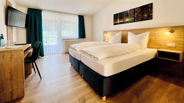 A hotel room with a bed, a desk, a chair, a TV, and a window with curtains. - Tanneneck (Löffingen)