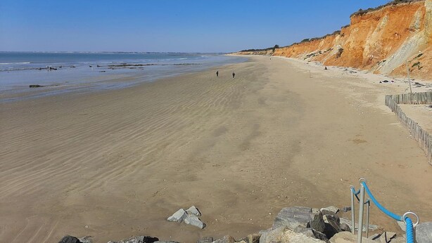 Une vaste plage de sable, avec une falaise rocheuse sur la droite et une étendue d’eau calme en arrière-plan.