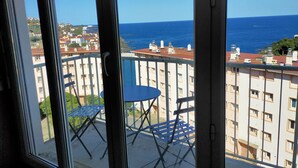 Un balcon avec une table et des chaises, donnant sur une zone côtière avec des bâtiments et la mer.