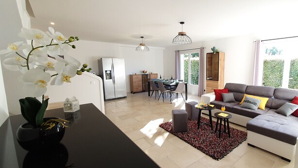 A modern living room with a grey sofa, a dining area with a wooden cabinet, and a kitchen with a refrigerator.