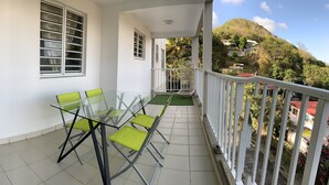 Een balkon met een glazen tafel en stoelen, een witte reling en uitzicht op een heuvel met gebouwen.