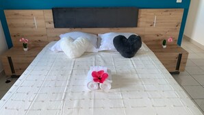 Een net opgemaakt bed met witte lakens, twee kussens, een opgerolde handdoek met een roze bloem en twee kleine potplanten op nachtkastjes.