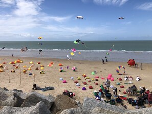Uma praia com muitos velejadores e bolas de praia, pessoas sentadas e de pé, e um primeiro plano rochoso.