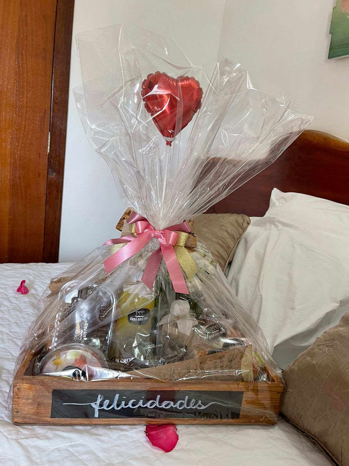Uma cesta de presentes sobre a cama, com cabeceira de madeira, contendo diversos itens e um balão em formato de coração amarrado com um laço rosa.