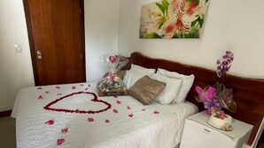 A neatly made bed with a heart-shaped arrangement of rose petals, a small bedside table with a vase of flowers, and a painting of flowers on the wall. - Pousada Buriti (Conceicao da Barra)