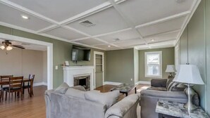 A living room with a fireplace, a television mounted above it, a dining area with chairs, and a sofa. - Riverview Townhouse II in Downtown Alexandria Bay (Alexandria Bay)