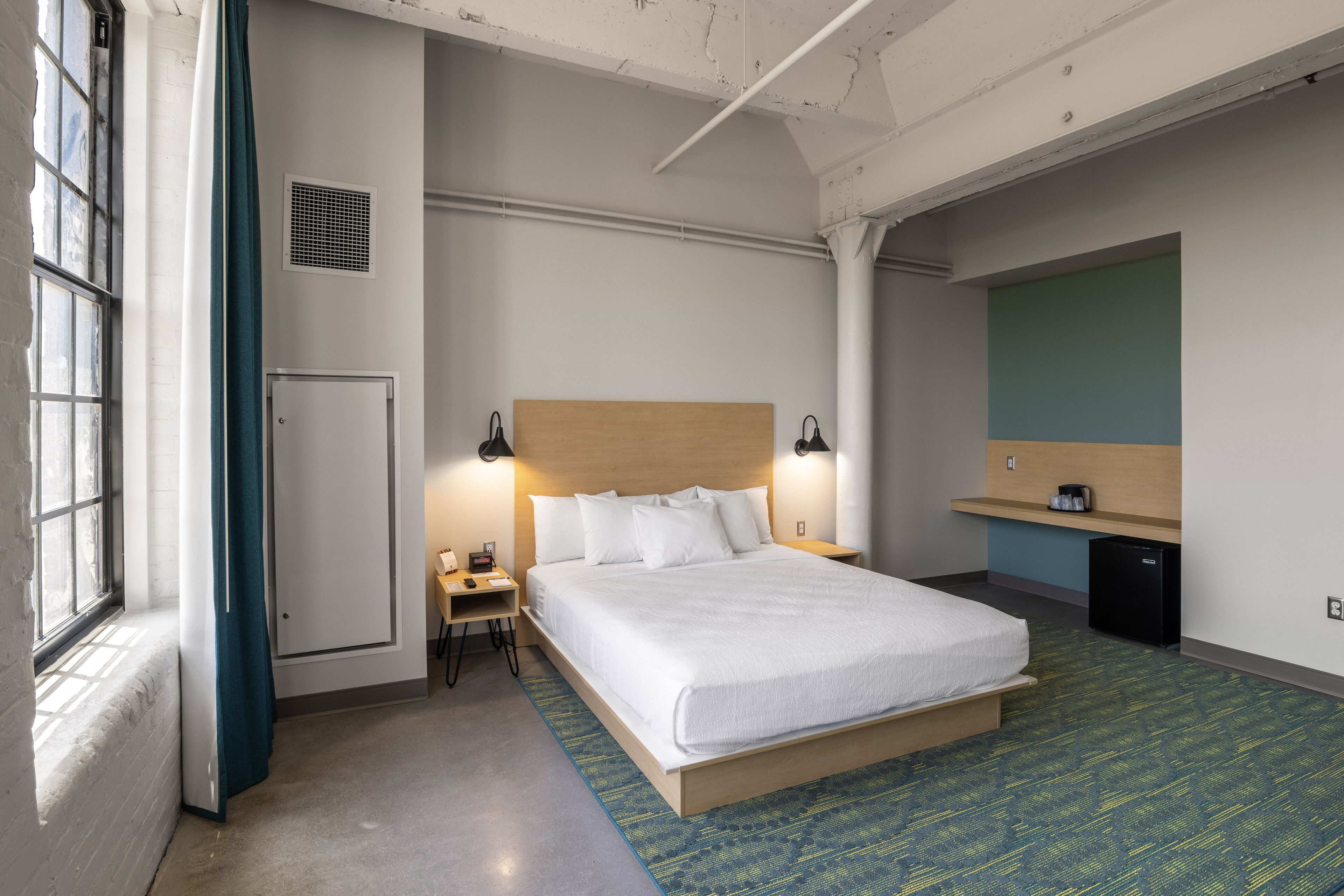 Habitación de hotel con una cama grande, un escritorio, una silla, una mesita, un espejo, un calentador y una ventana con cortinas.