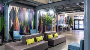 A spacious lobby with a high ceiling, exposed beams, and a colorful geometric wall design. There are several seating areas with cushions and a staircase leading to an upper level. - The Warehouse Hotel at Champion Mill (Hamilton)