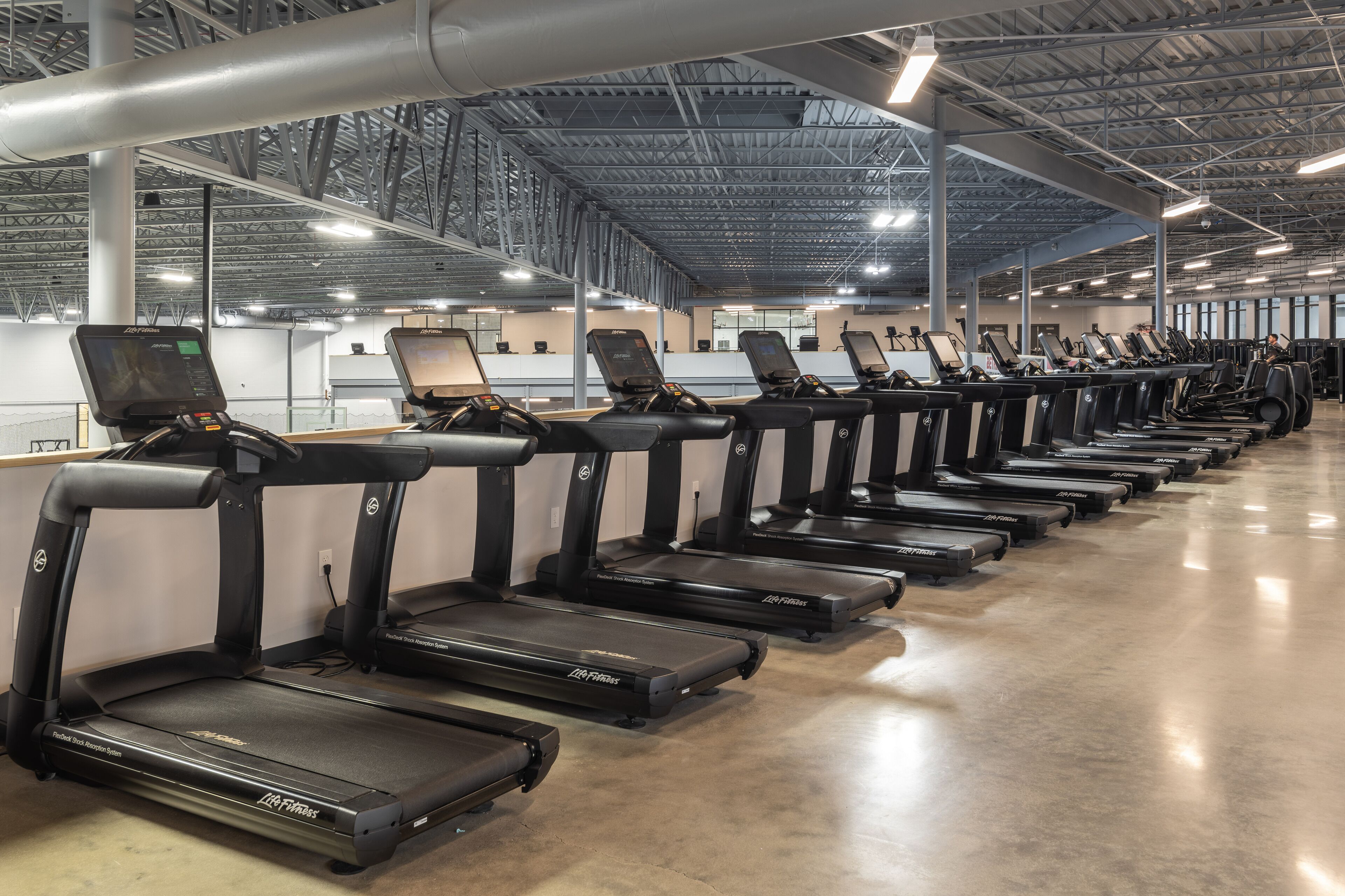 A row of treadmills in a gym with a high ceiling and visible structural beams.