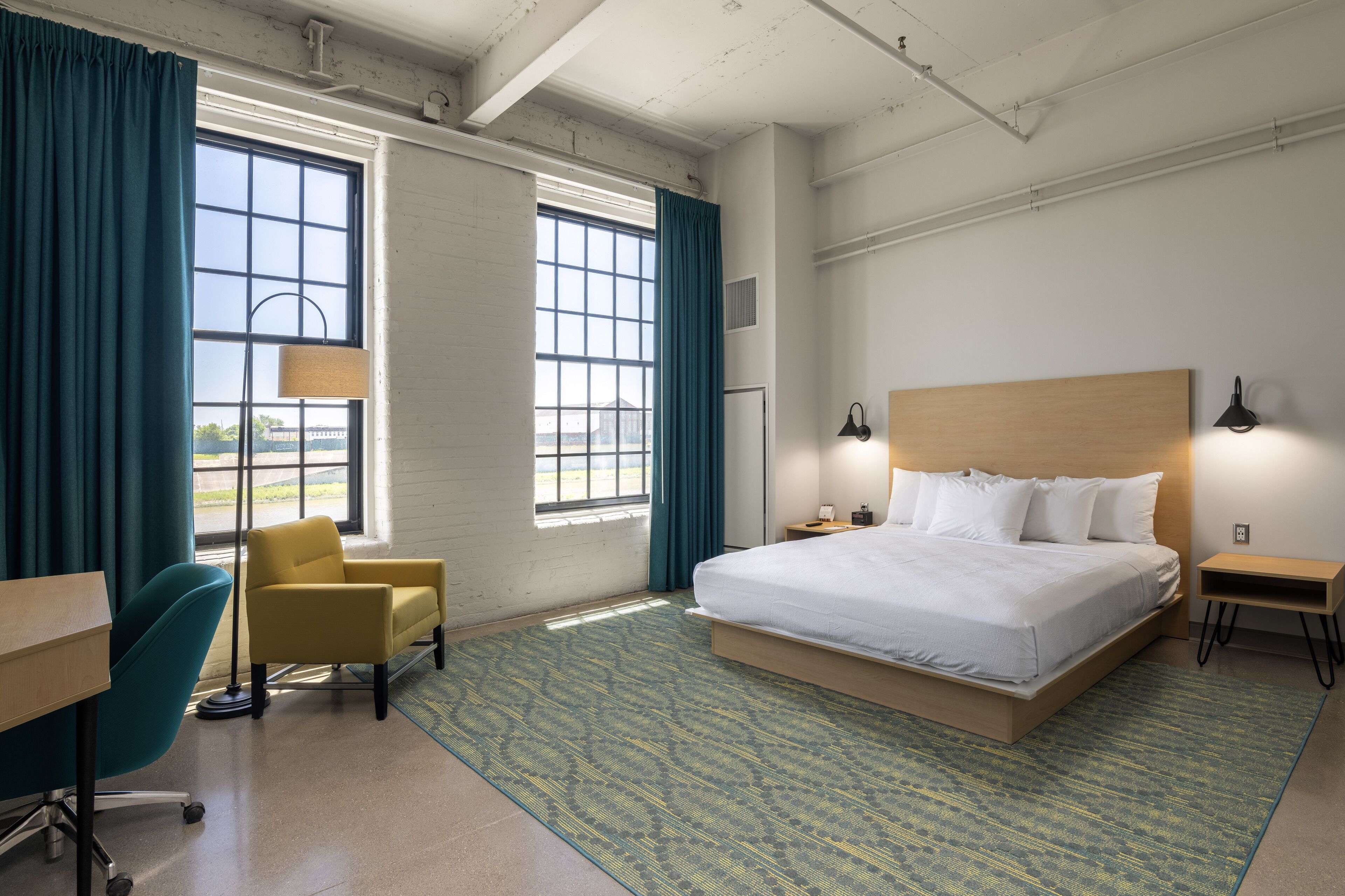 Una habitación de hotel con una cama grande, un escritorio, una silla y dos ventanas con cortinas.