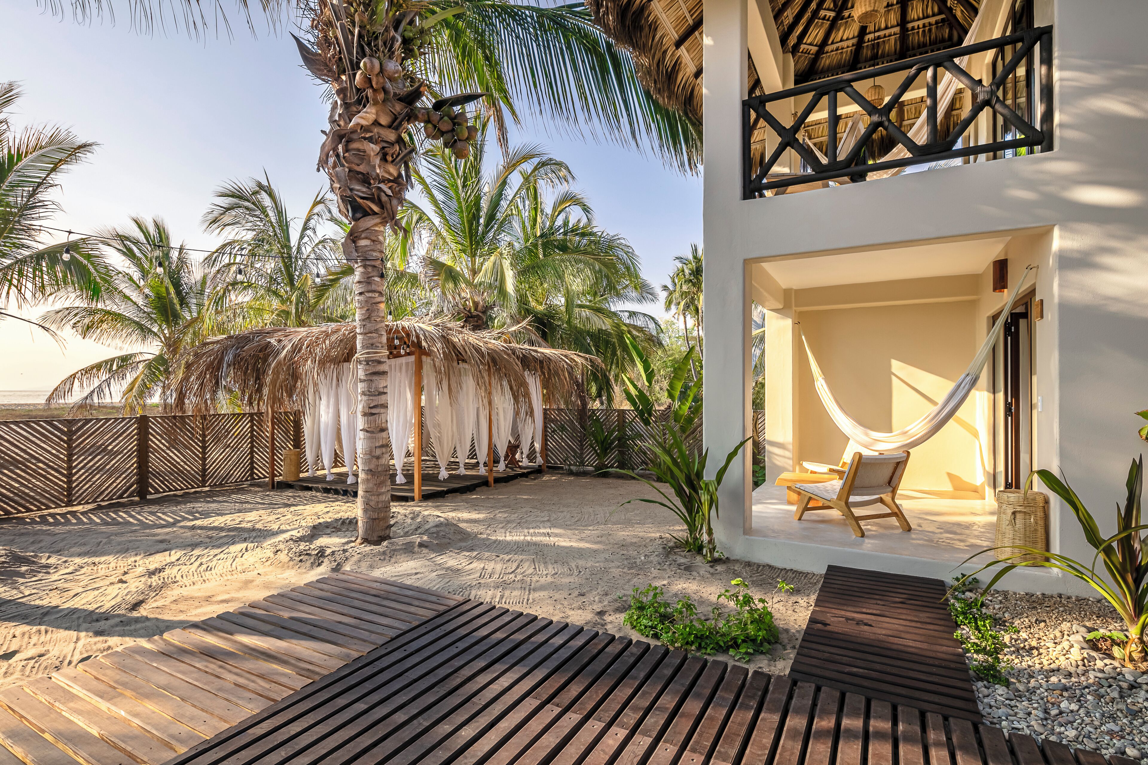 A beachside villa with a hammock, wooden deck, palm trees, and a thatched-roof structure.