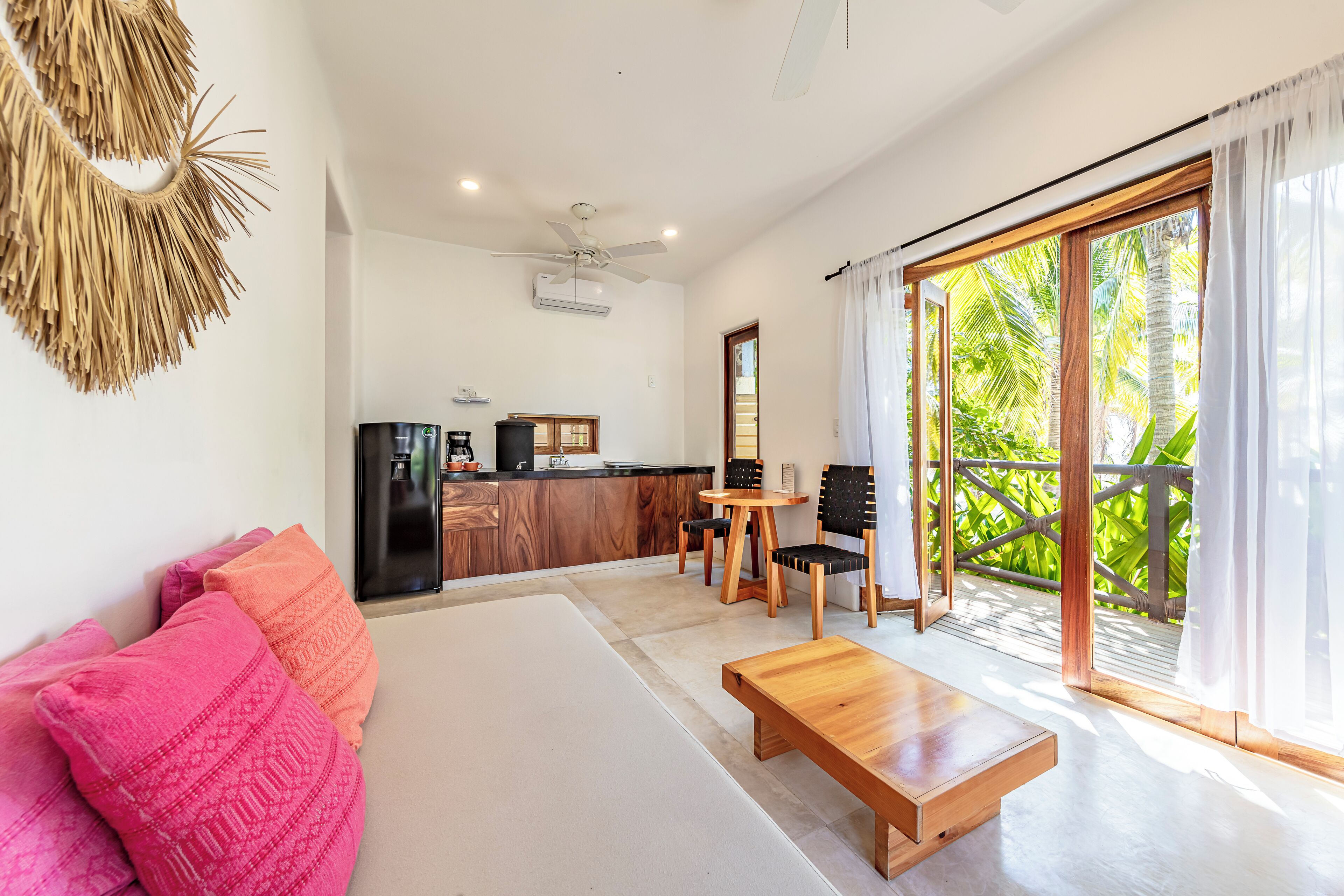 Un espacio habitable moderno con sofá, mesa de centro y comedor. Se ve una cocina al fondo, y una puerta corrediza de vidrio da a un balcón con plantas.