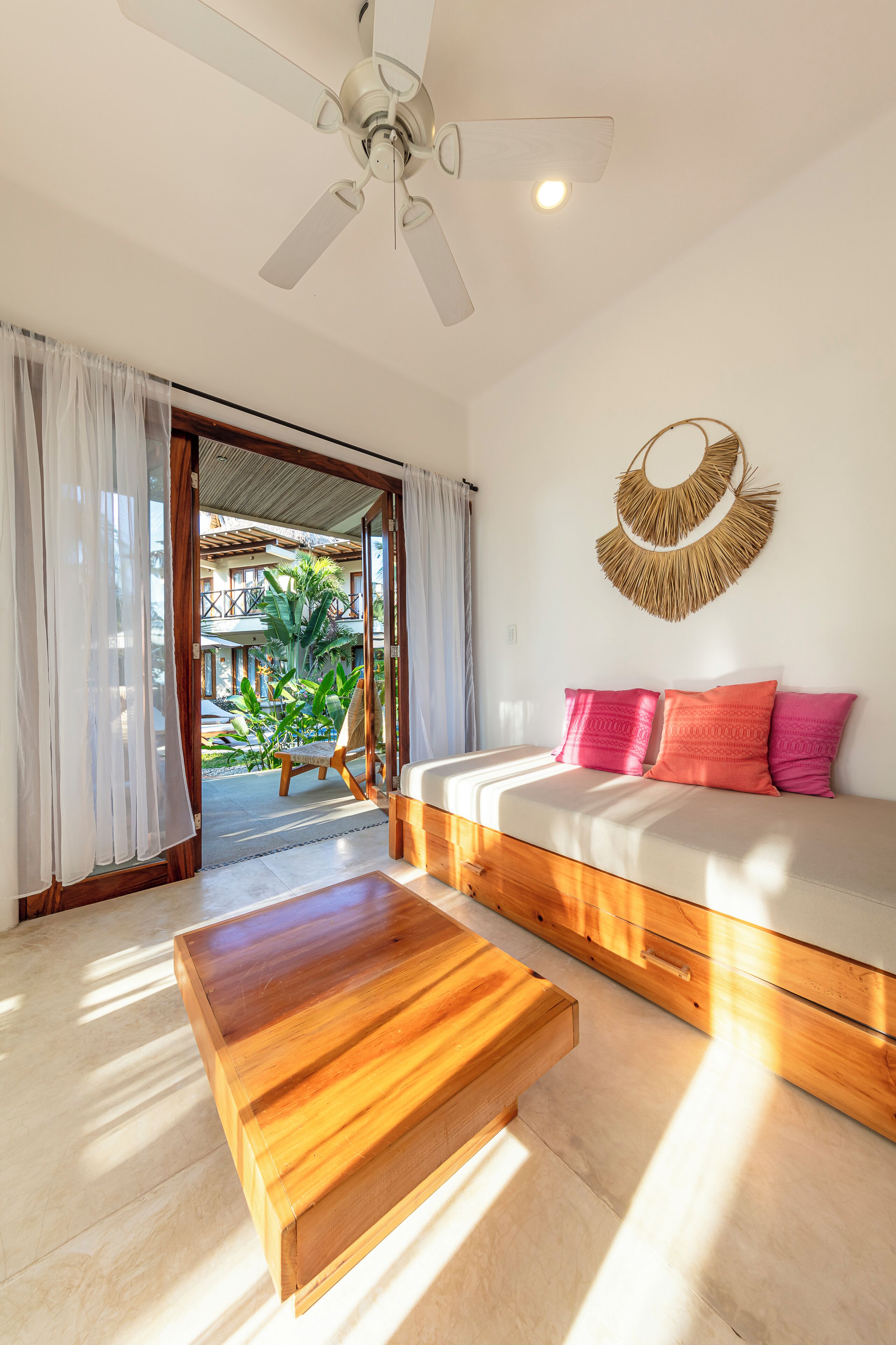 A room with a bed, a wooden coffee table, a ceiling fan, and a view of an outdoor area with plants.