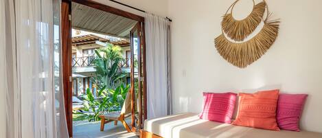 A room with a bed, a wooden coffee table, a ceiling fan, and a view of an outdoor area with plants.