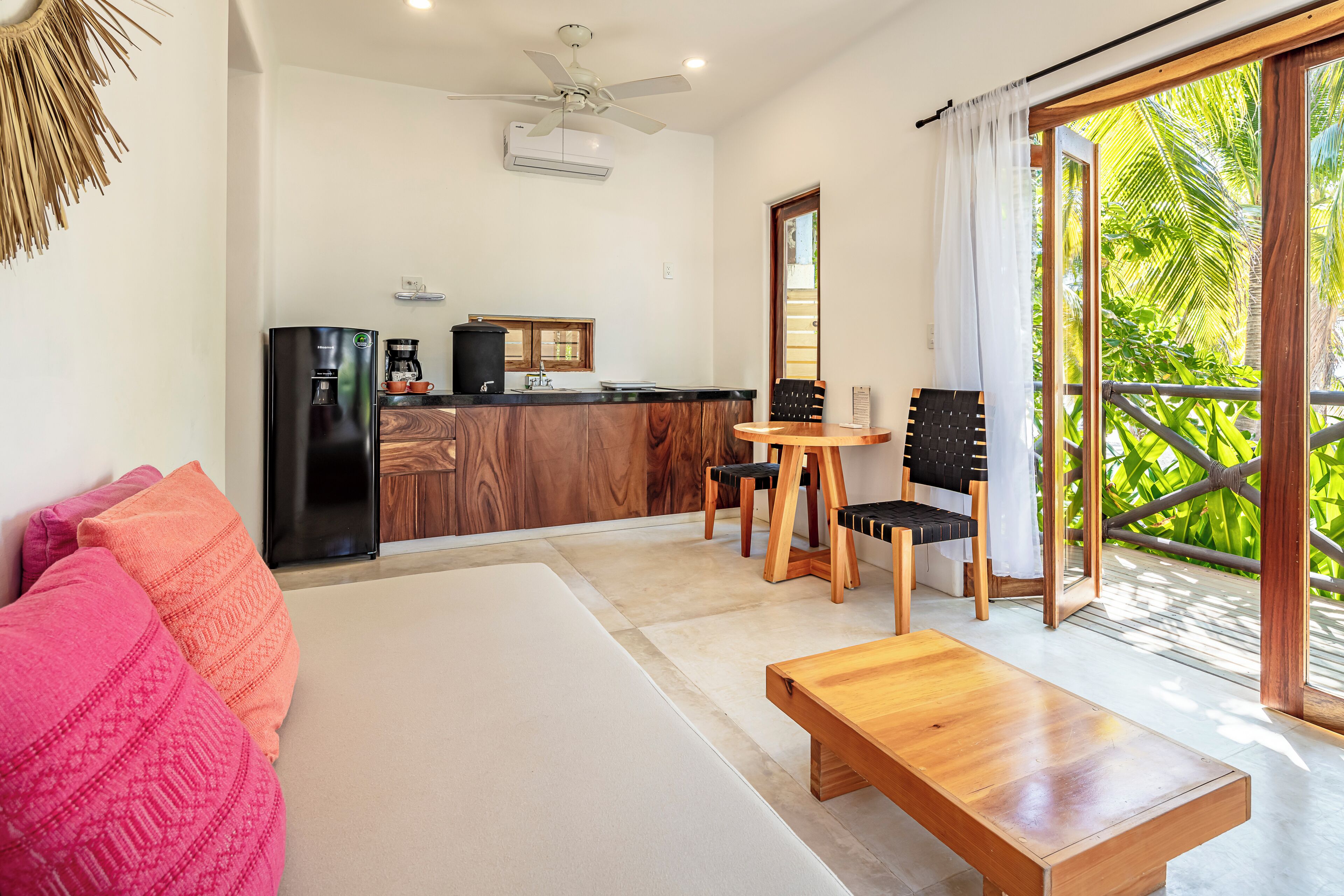 A modern room with a bed, a wooden bench, a dining table with chairs, a kitchen area, and a balcony with a view of greenery.