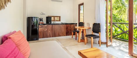 A modern room with a bed, a wooden bench, a dining table with chairs, a kitchen area, and a balcony with a view of greenery.