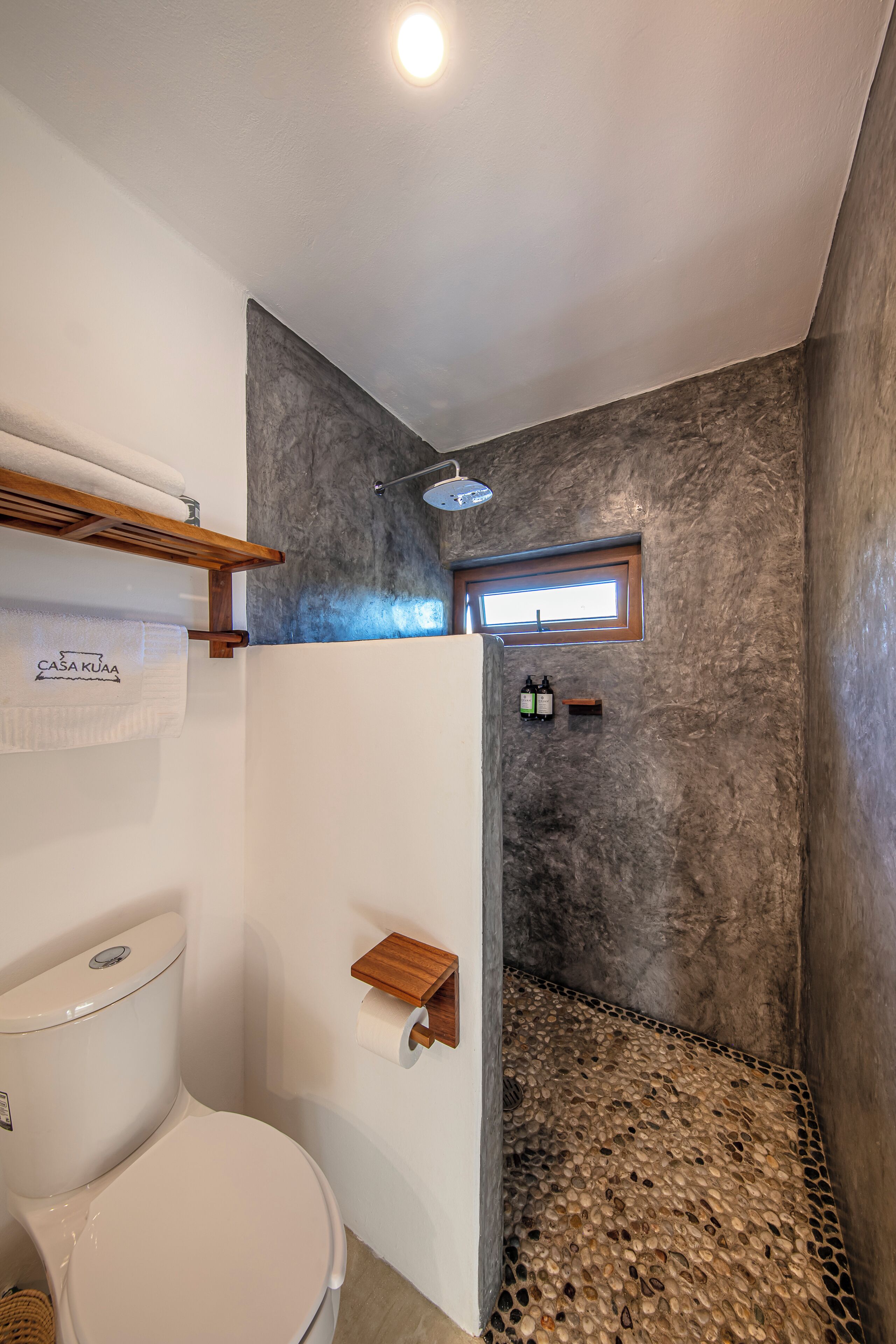 A bathroom with a white toilet, a towel rack with a towel, a shower area with a textured wall, and a small window.
