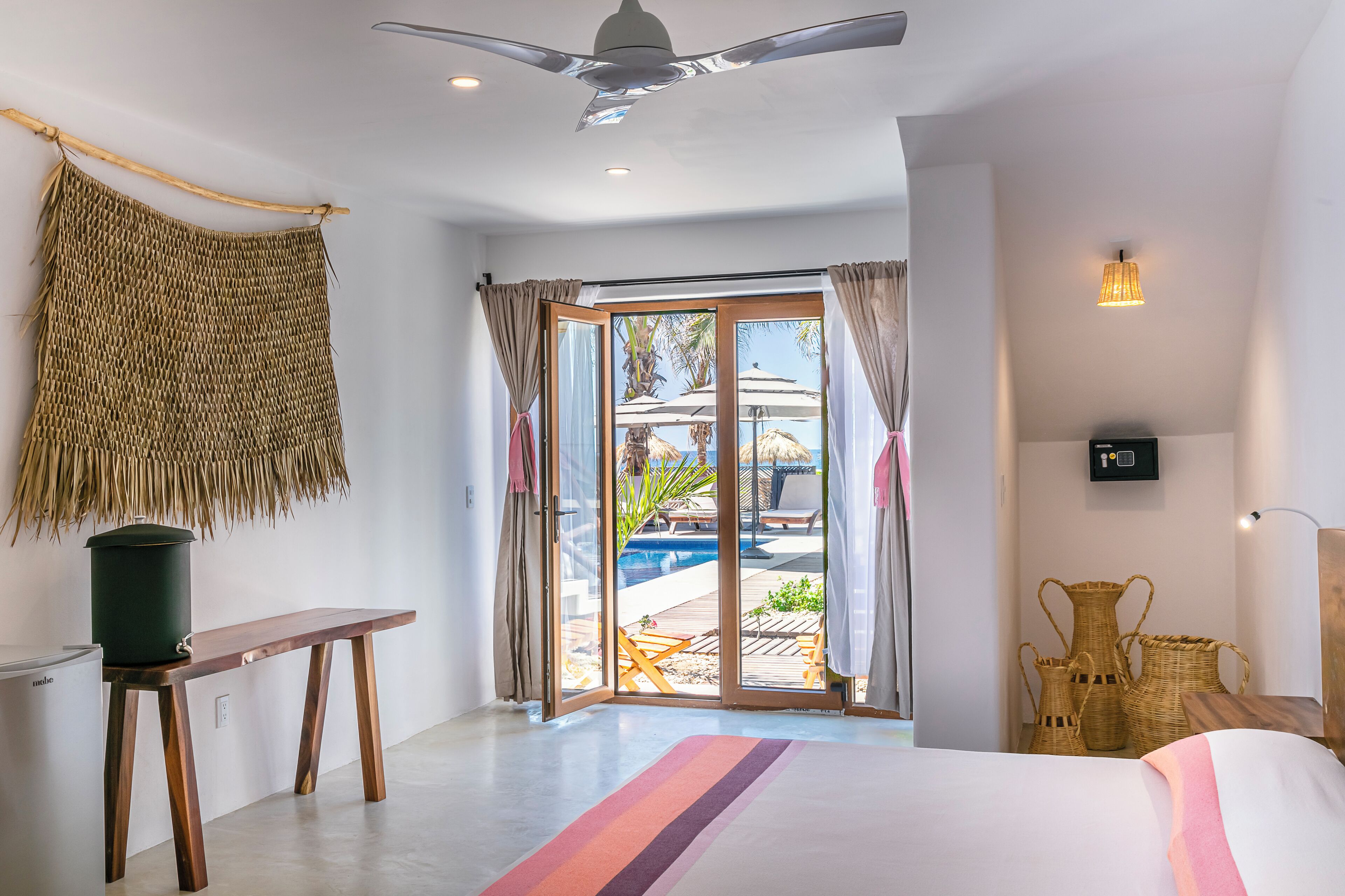 A bedroom with a bed, a wooden bench, a woven wall hanging, and a view of an outdoor pool area.