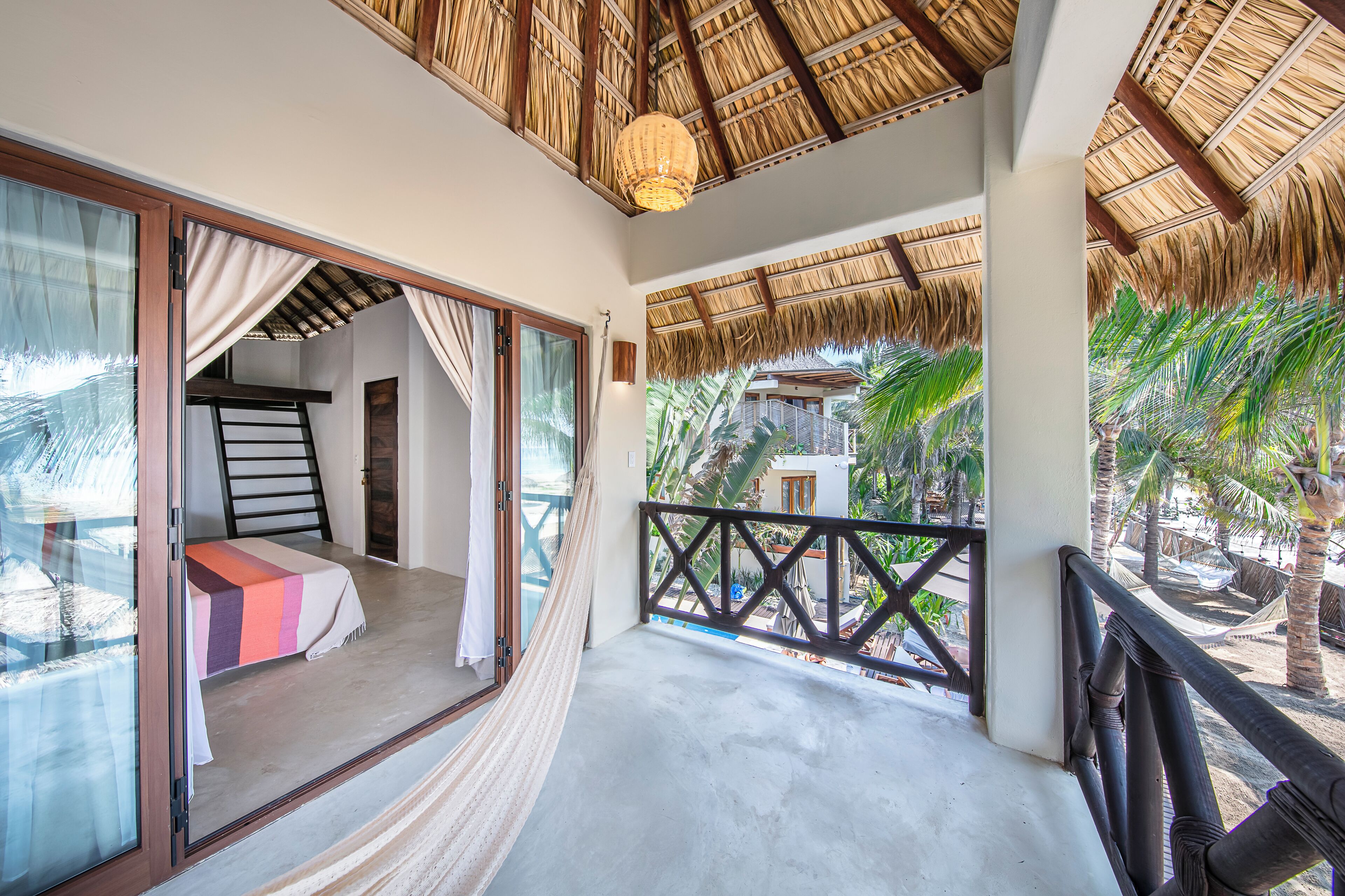 A balcony with a hammock, a bed with a striped blanket, and a view of palm trees.