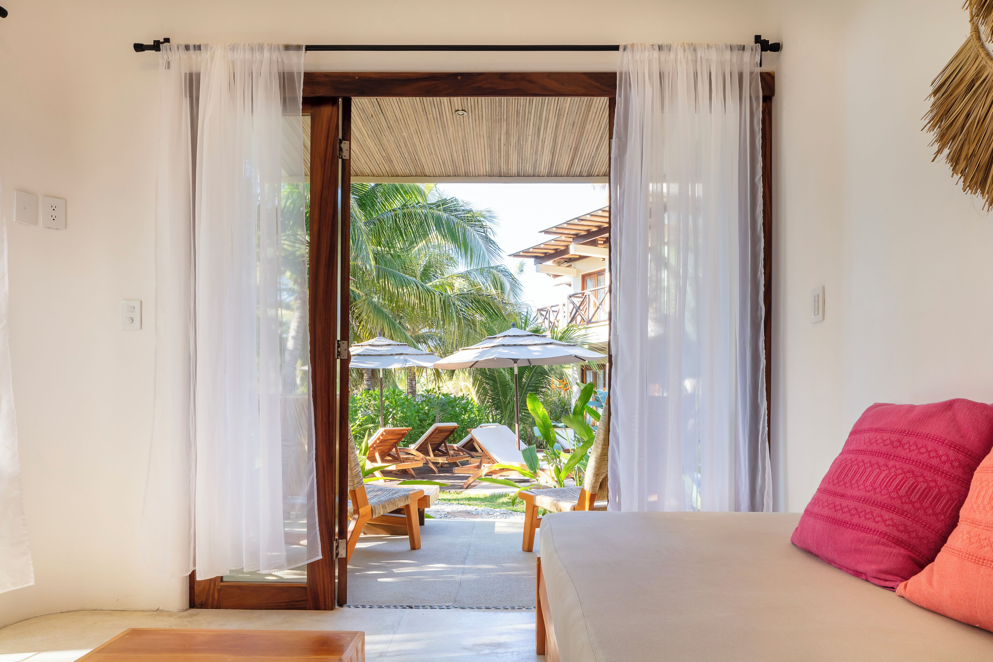 A room with a bed, a view of a pool area with lounge chairs and umbrellas, and a tropical garden.