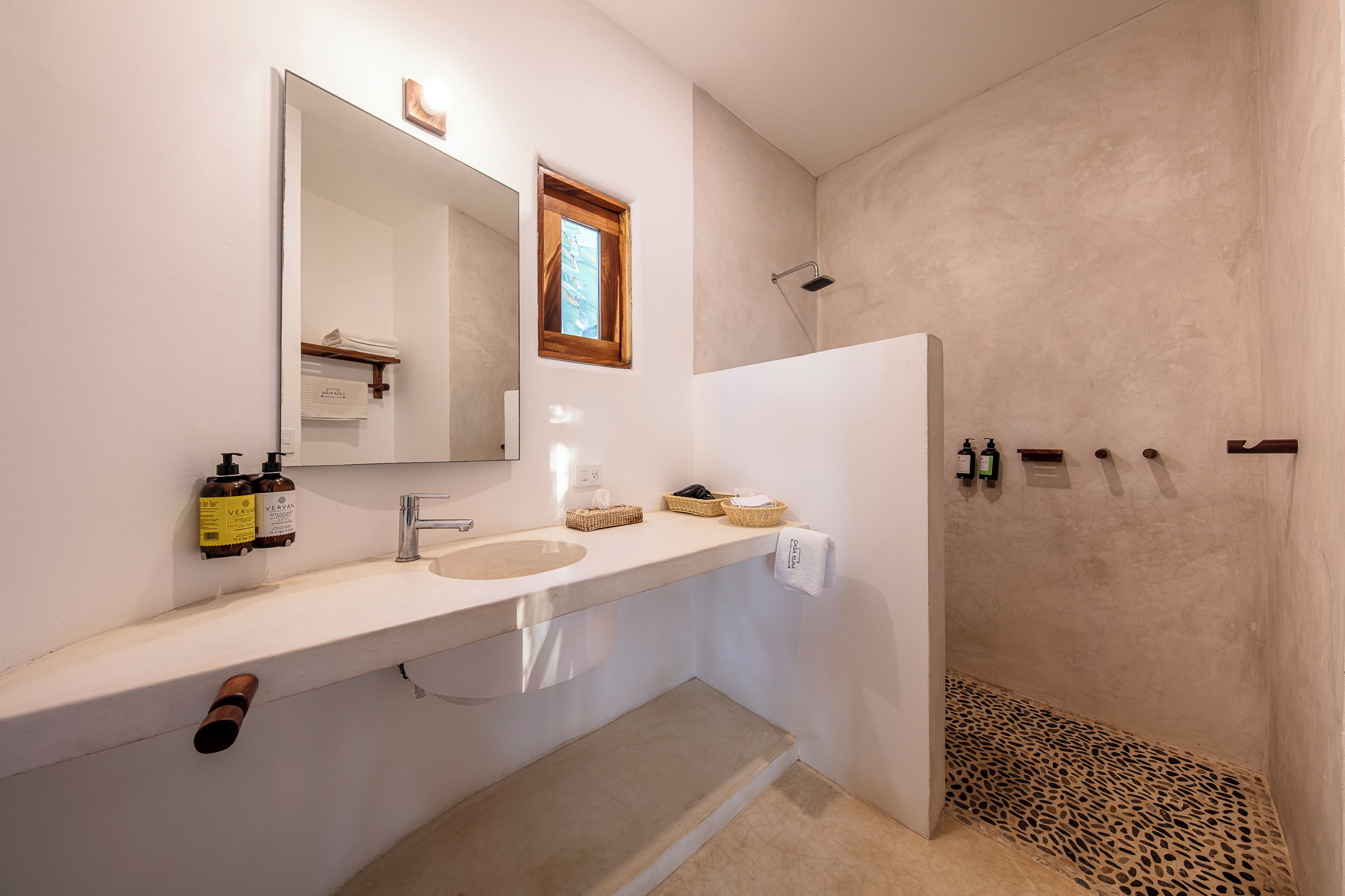 A modern bathroom with a large mirror, a sink, and a shower. There are toiletries on the counter and a towel hanging on the wall.