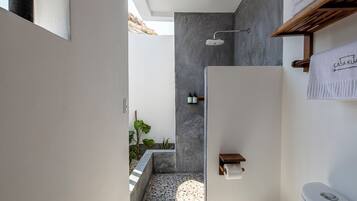 Un baño moderno con un inodoro blanco, una ducha y un lavamanos pequeño. Hay un perchero con una toalla colgando, y una cesta en el suelo.