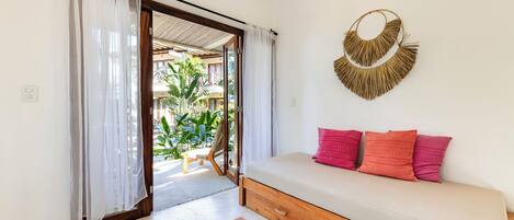 A room with a wooden sofa, a woven wall hanging, and a view of a balcony with plants.