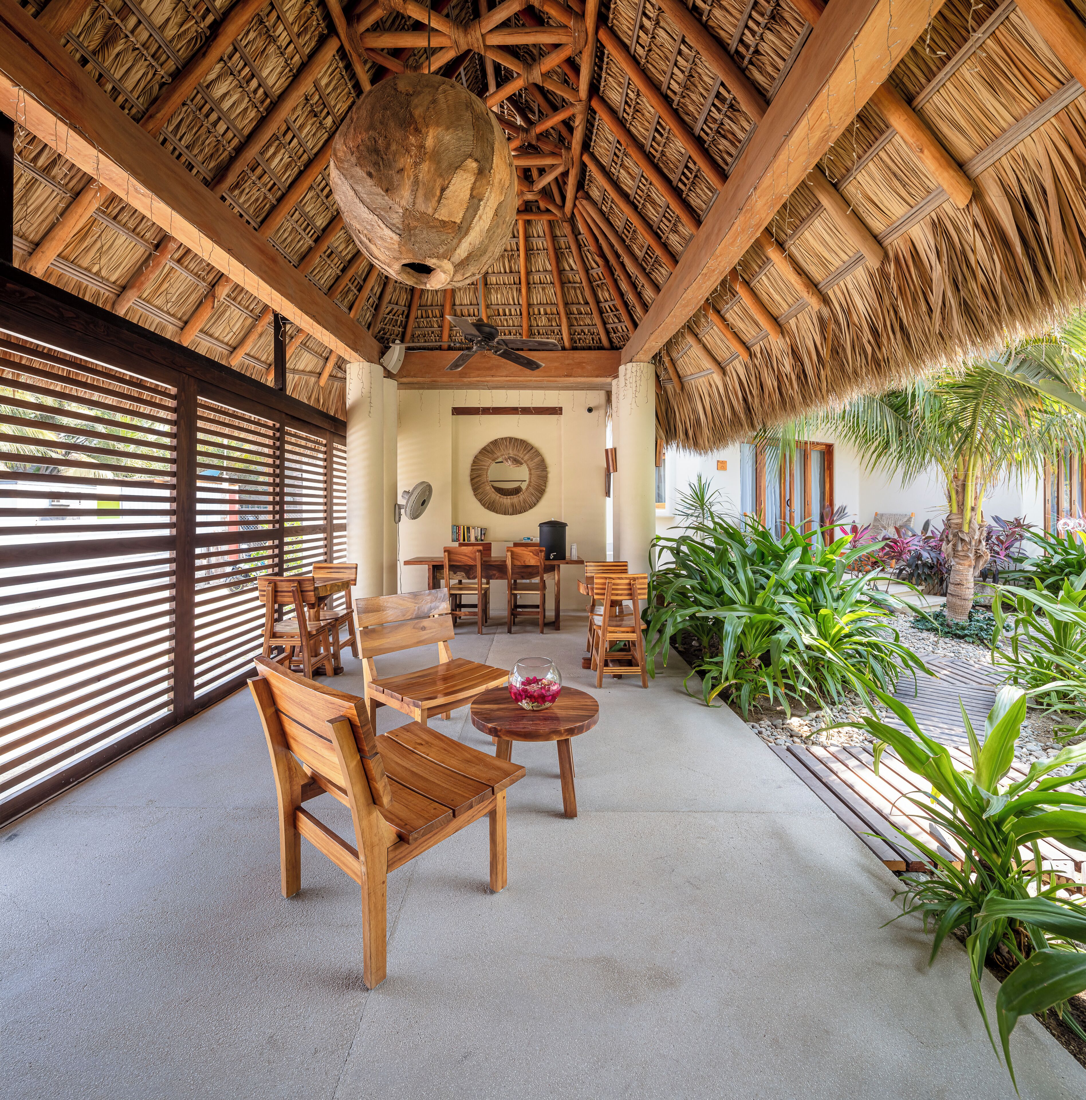 Un área de estar al aire libre con techo, muebles de madera, ventilador de techo y vista a un jardín con plantas tropicales.