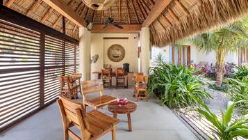 Un área de estar al aire libre con techo, muebles de madera, ventilador de techo y vista a un jardín con plantas tropicales.