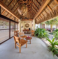 Un área de estar al aire libre con techo, muebles de madera, ventilador de techo y vista a un jardín con plantas tropicales.