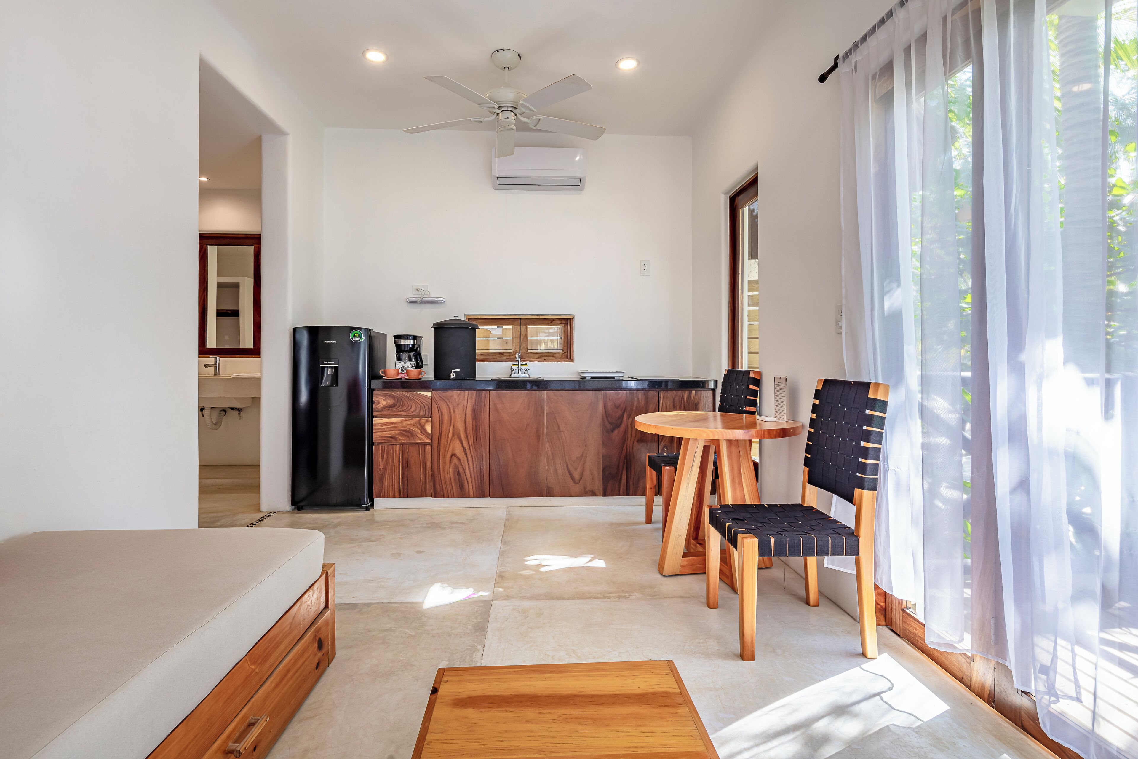 A compact room with a bed, a dining table with chairs, a kitchen area with a refrigerator, and a ceiling fan.