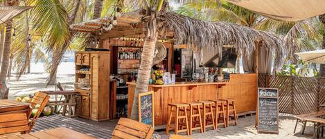 Un bar situé en bord de mer, avec des meubles en bois, un tableau noir servant de menu, et un décor tropical agrémenté de palmiers.